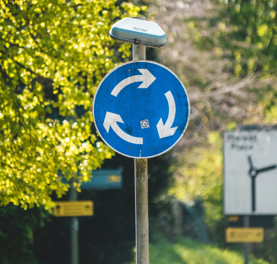 Ein blaues Verkehrsschild mit weißen Pfeilen, das einen Kreisverkehr symbolisiert. Im Hintergrund ist eine grüne Landschaft mit Bäumen und Wegweisern zu sehen. Das Schild könnte symbolisch für die Kreislaufwirtschaft stehen, die auf Wiederverwendung und Recycling basiert.
