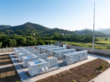 Drohnenaufnahme einer großen Batteriespeicher-Anlage im Freien. Mehrere Reihen weißer Batterie-Container stehen auf einem Betonfundament, umgeben von grüner Landschaft und Hügeln unter blauem Himmel.