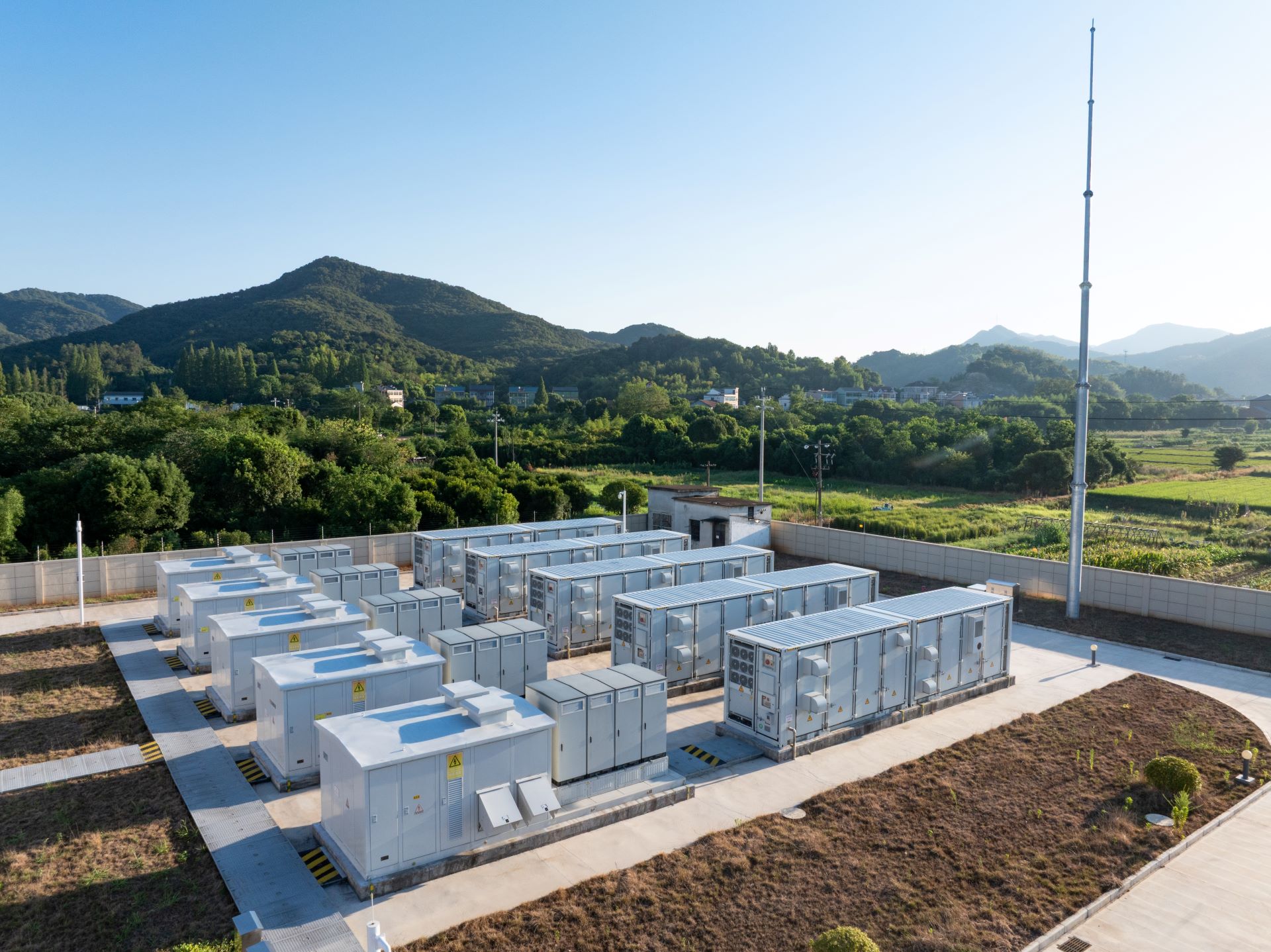 Drohnenaufnahme einer großen Batteriespeicher-Anlage im Freien. Mehrere Reihen weißer Batterie-Container stehen auf einem Betonfundament, umgeben von grüner Landschaft und Hügeln unter blauem Himmel.