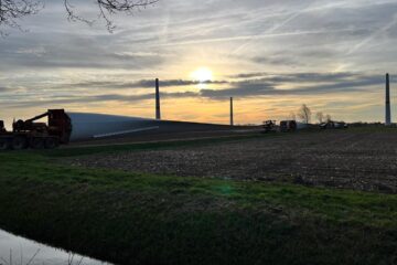Stimmungsvolle Abenddämmerung auf dem Gelände des Bürgerwindparks Wagenfeld-Süd: Im Vordergrund liegt ein langes weißes Rotorblatt auf einem Schwertransporter. Im Hintergrund sind mehrere bereits errichtete Windkrafttürme als Silhouetten vor einem orange-goldenen Sonnenuntergangshimmel zu sehen. Ein Entwässerungsgraben verläuft im Vordergrund entlang eines Ackers.