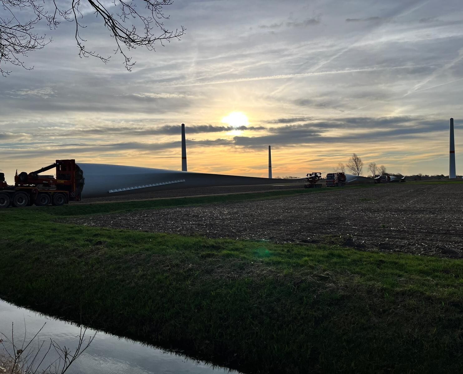 Stimmungsvolle Abenddämmerung auf dem Gelände des Bürgerwindparks Wagenfeld-Süd: Im Vordergrund liegt ein langes weißes Rotorblatt auf einem Schwertransporter. Im Hintergrund sind mehrere bereits errichtete Windkrafttürme als Silhouetten vor einem orange-goldenen Sonnenuntergangshimmel zu sehen. Ein Entwässerungsgraben verläuft im Vordergrund entlang eines Ackers.