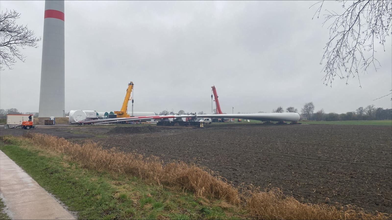 Tageslichtaufnahme vom Windparkgelände: Mehrere lange weiße Rotorblätter liegen auf dem Boden. Im Hintergrund steht ein grau-weißer Windkraftturm mit rotem Streifen. Zwei Kräne (gelb und rot) sind beim Abladen im Einsatz.
