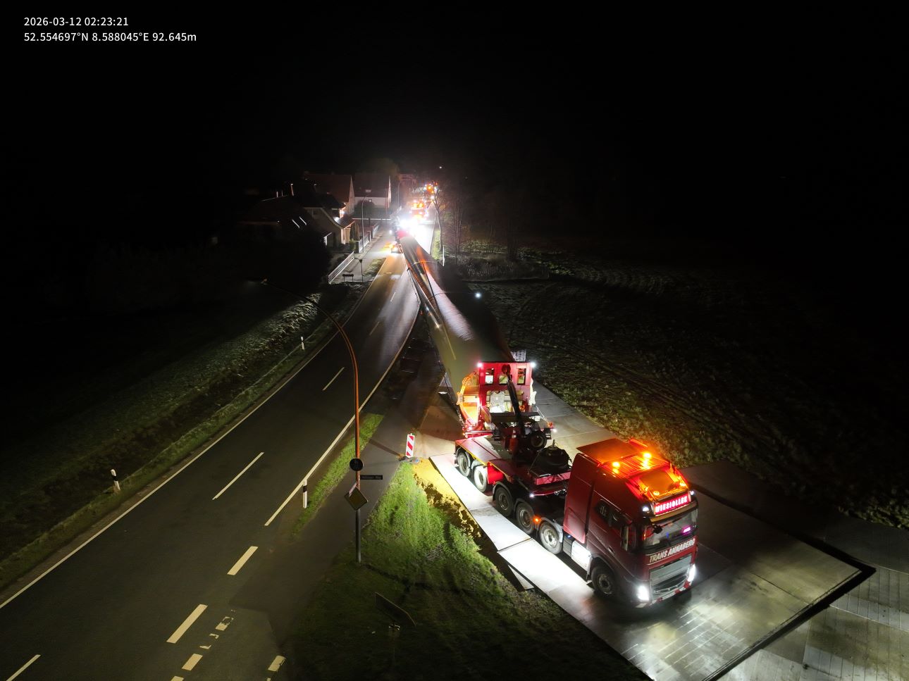 Drohnenaufnahme bei Nacht: Ein Schwertransporter mit einem langen Rotorblatt fährt durch eine beleuchtete Landstraße in einer Ortschaft. GPS-Koordinaten zeigen den Standort nahe Wagenfeld, Aufnahmezeit 02:23 Uhr am 12. März 2026.