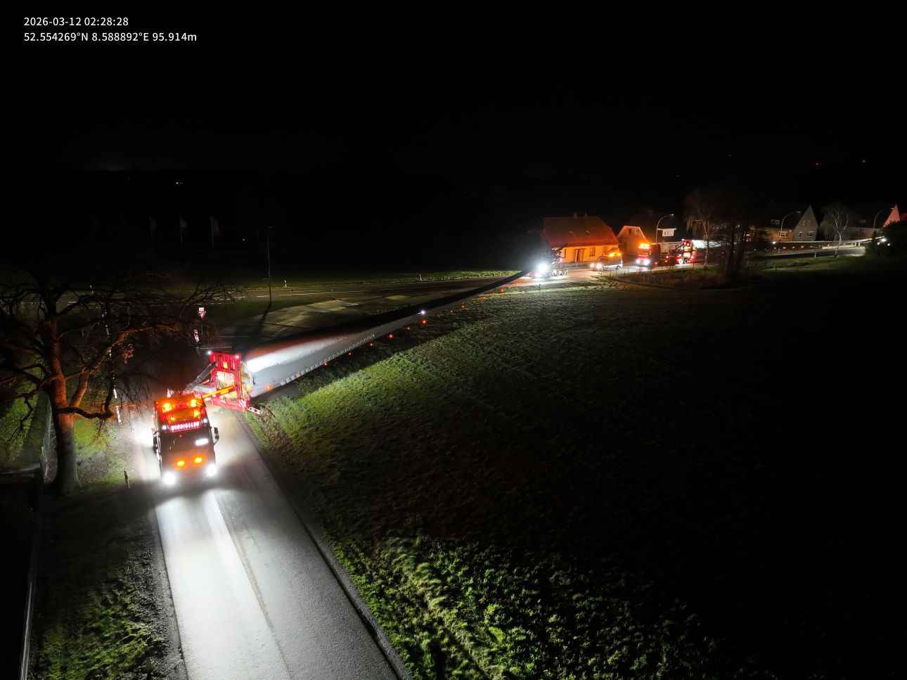 Drohnenaufnahme bei Nacht: Ein Schwertransporter mit einem langen Rotorblatt fährt auf einer schmalen Landstraße zwischen Feldern und Wohnhäusern. Begleitfahrzeuge mit Warnlichtern sind sichtbar. Aufnahmezeit 02:28 Uhr am 12. März 2026.