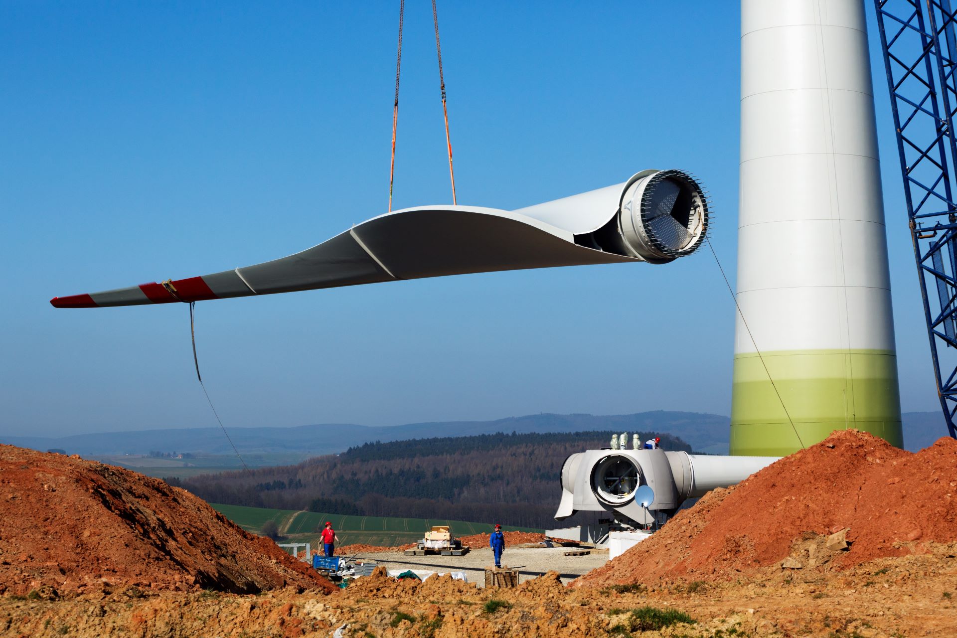 Montage eines Windkraftanlagen-Rotorblatts auf einer Baustelle: Ein Kran hebt ein großes weißes Rotorblatt in die Höhe, im Hintergrund steht der bereits aufgerichtete weiß-grüne Turm der Windkraftanlage sowie die Gondel am Boden. Mehrere Arbeiter sind auf dem Baugelände sichtbar.