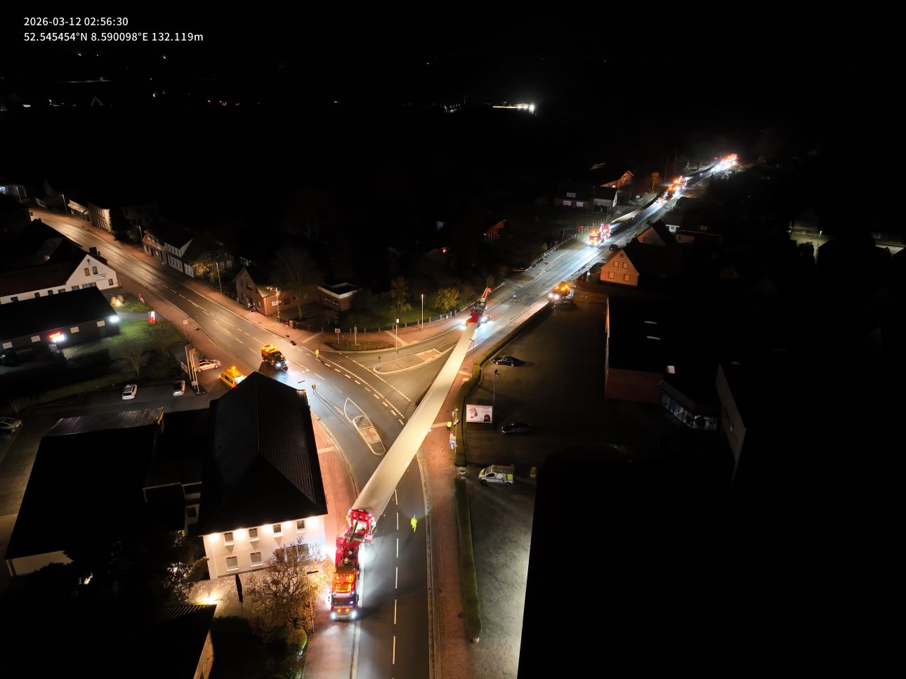 Drohnenaufnahme bei Nacht: Ein Schwertransporter mit Rotorblatt durchfährt eine beleuchtete Kreuzung in einer Ortschaft. Begleitfahrzeuge sichern den Konvoi ab. Aufnahmezeit 02:56 Uhr am 12. März 2026.